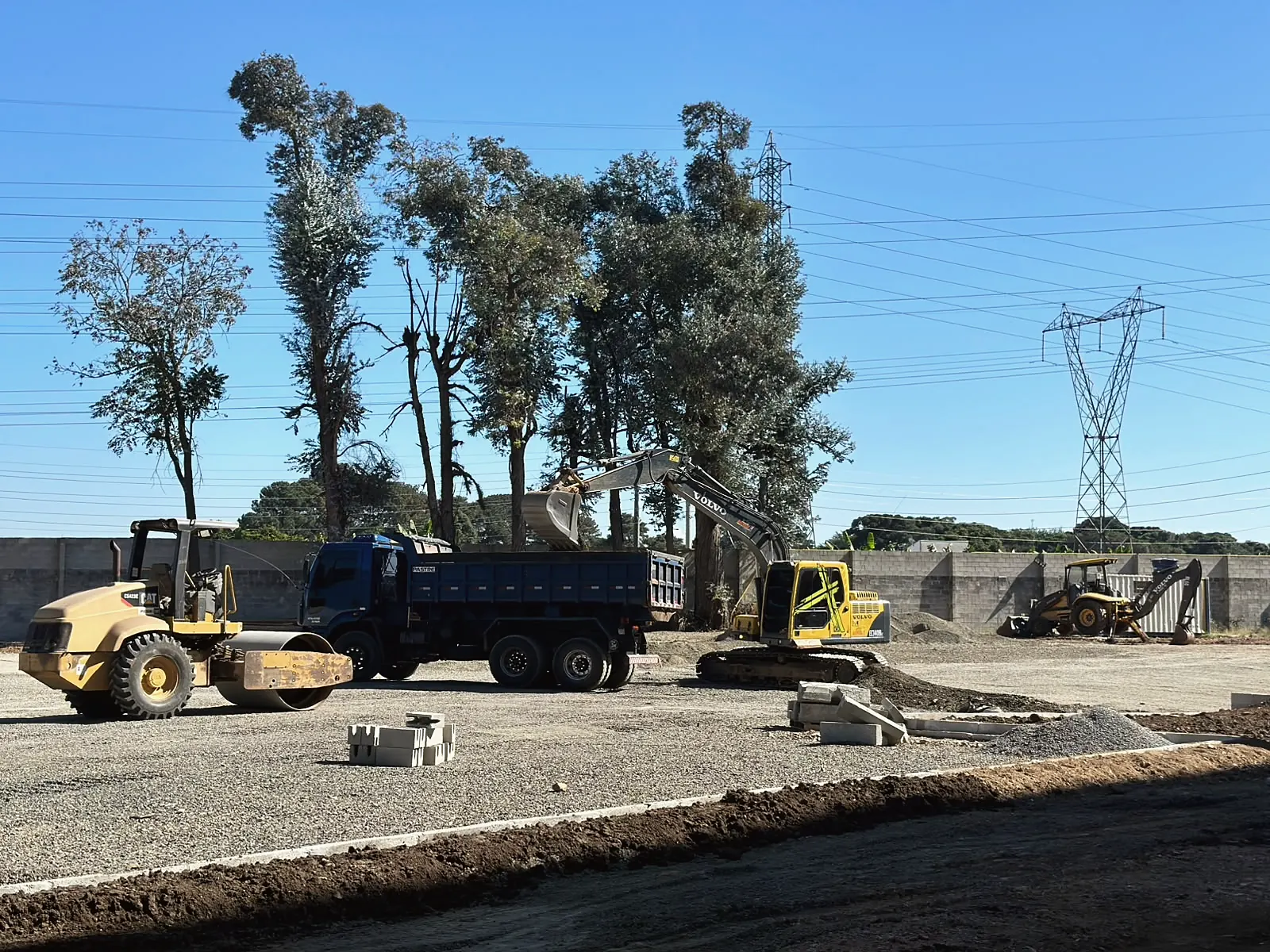 Rolo compactador, caminhão basculante e escavadeira trabalhando juntos na terraplanagem de um terreno amplo