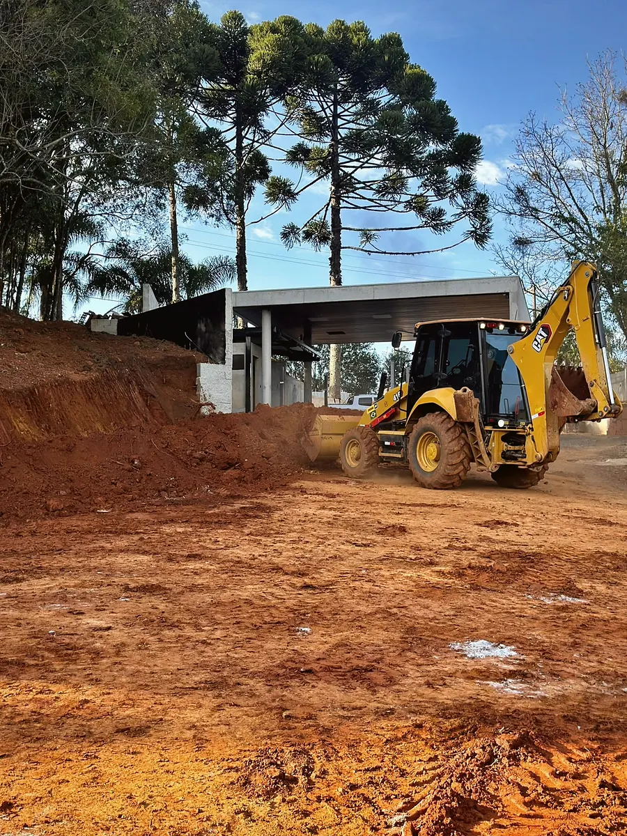 Retroescavadeira trabalhando em terreno de terra vermelha com araucárias ao fundo